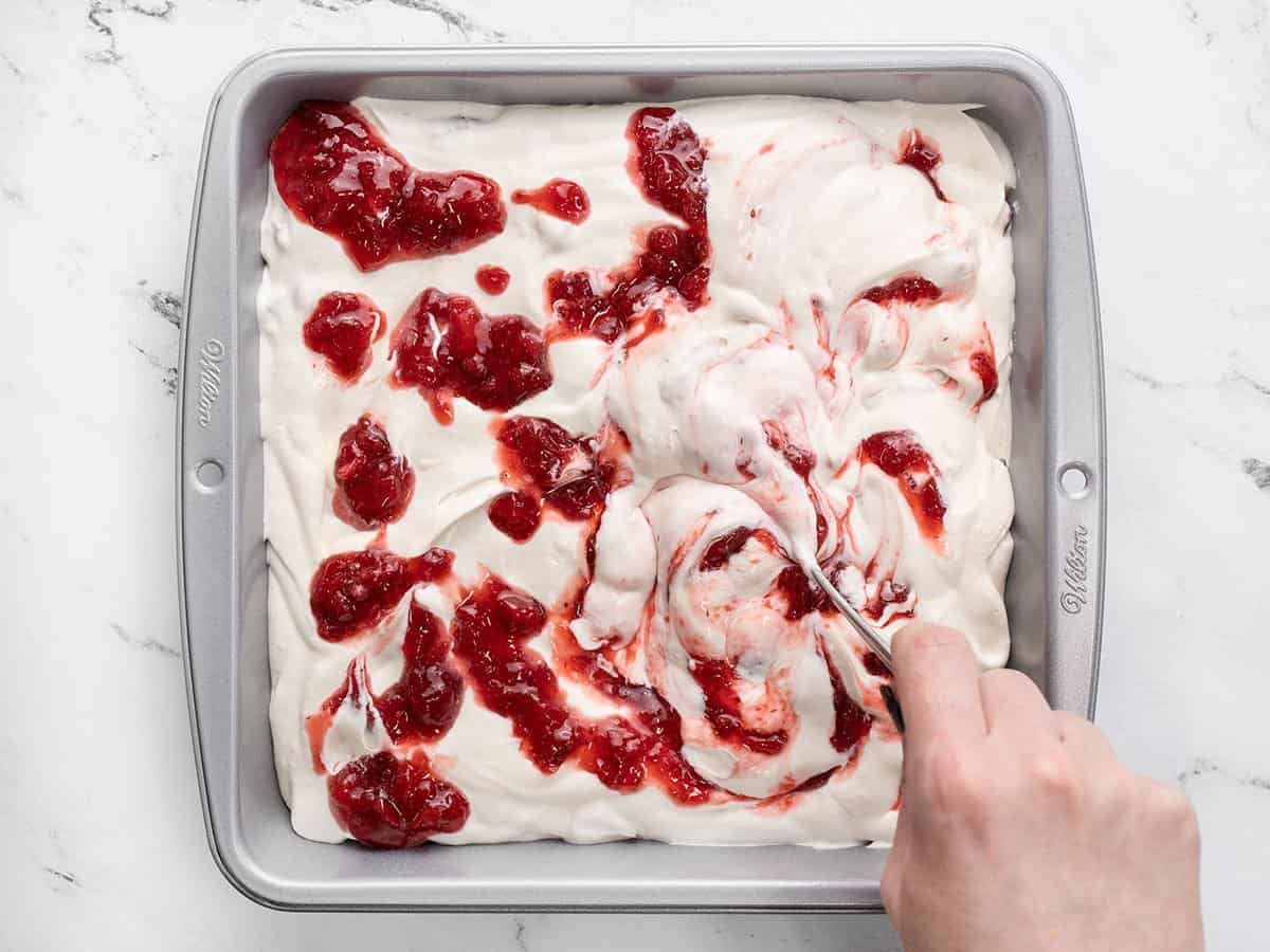 Strawberry syrup is being swirled into the ice cream base in a freezer-safe container.