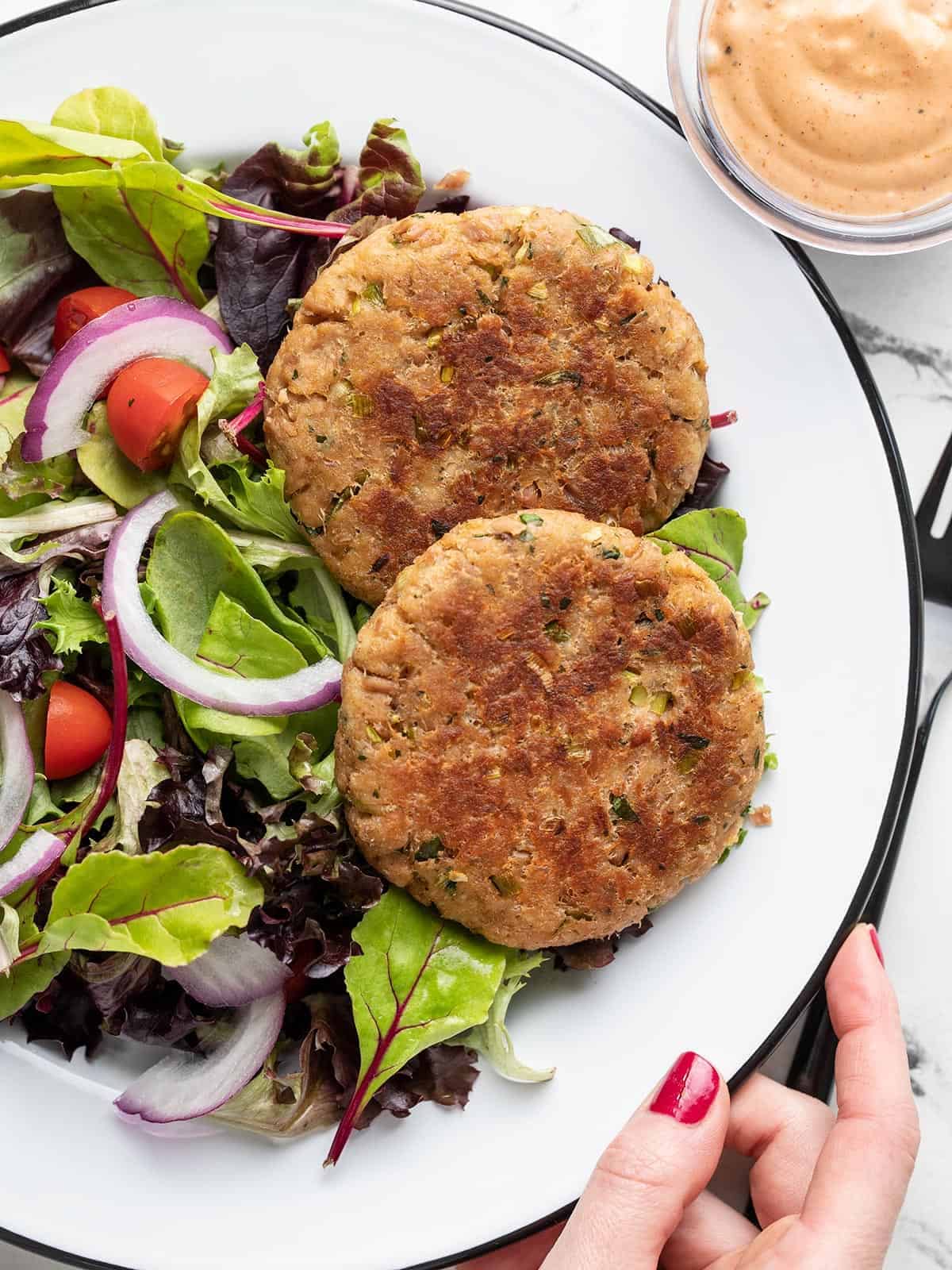 Two tuna patties on a plate with greens, a hand holding the side of the plate.