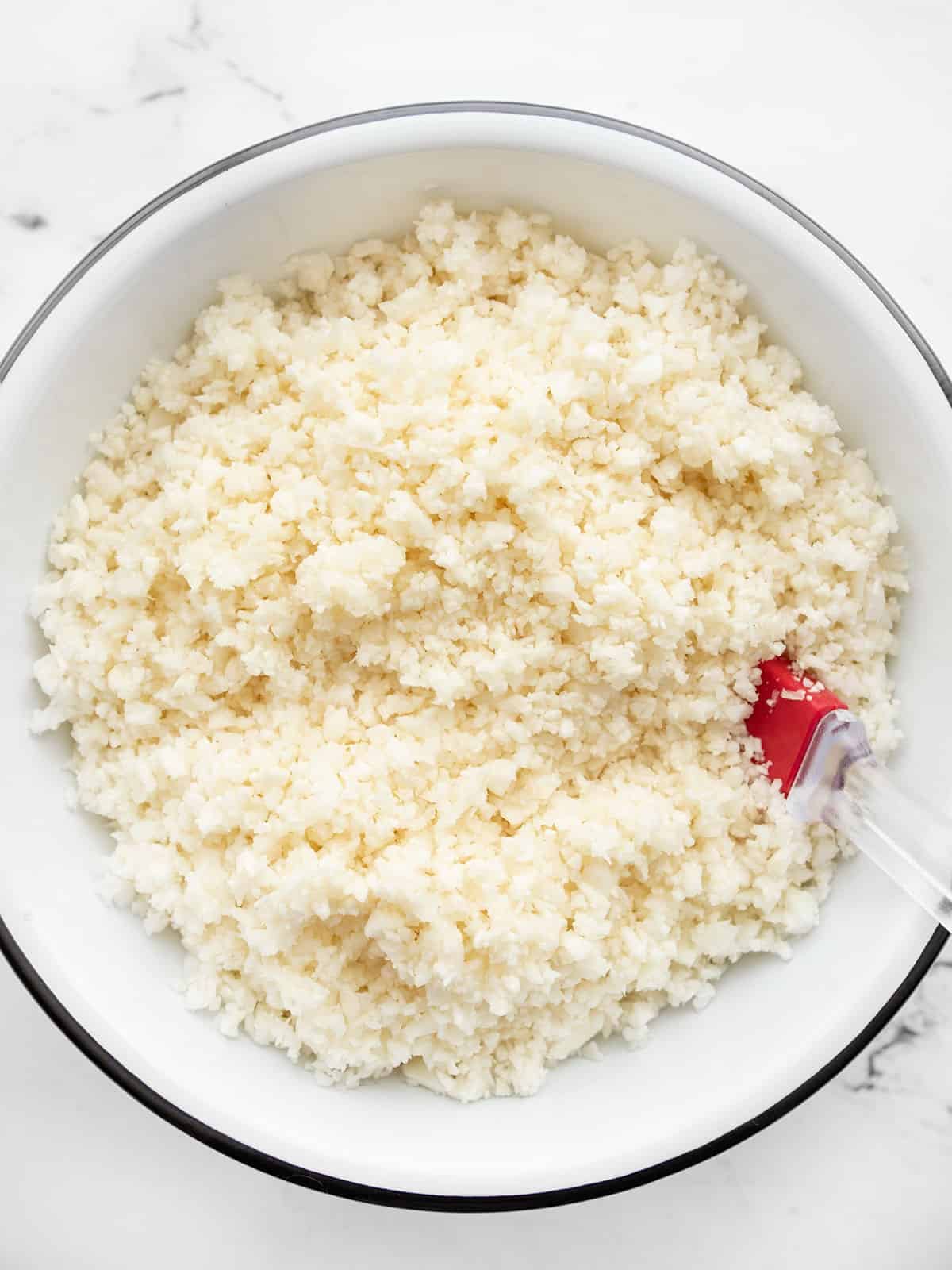 Overhead view of a bowl of riced cauliflower with a red spatula in the side