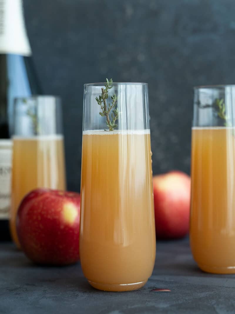 three apple cider mimosas with apples and a bottle of wine in the back