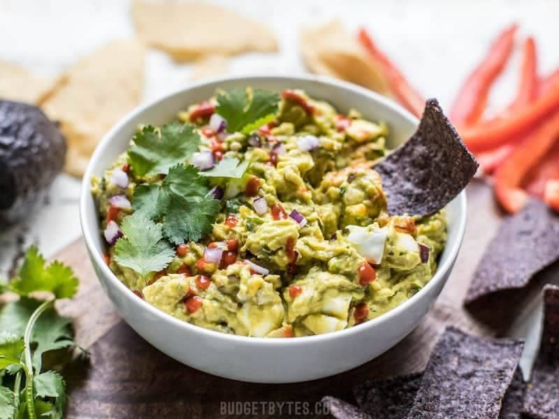 Side view of a bowl of loaded guacamole with a chip dipped in it 