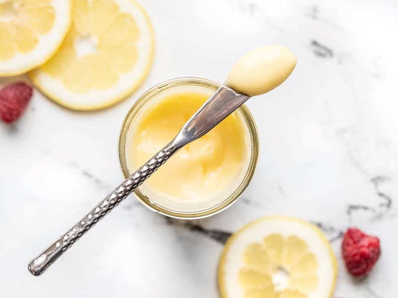 Refrigerated lemon curd on a small butter knife set across the mouth of a jar, lemon slices on the side.