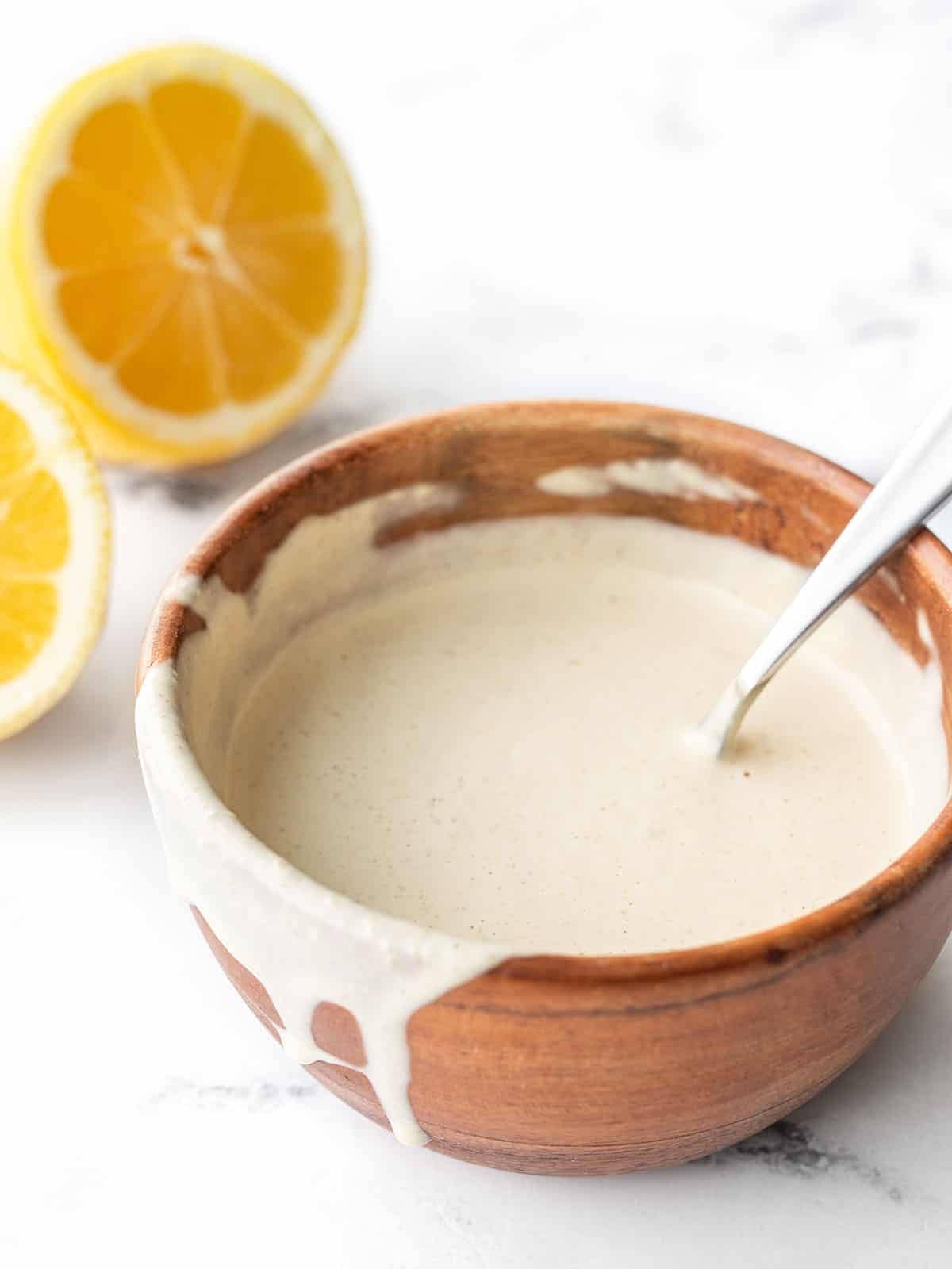 Lemon tahini dressing in a wooden bowl dripping over the side, a cut lemon in the back