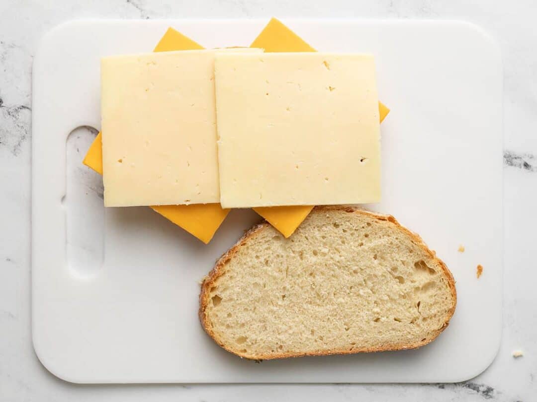 Cheese slices for air fryer grilled cheese on top of one slice of bread with a second slice of bread next to it, on a cutting board.