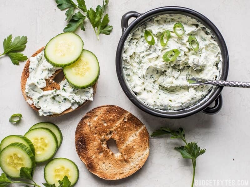 A bowl of Scallion Herb Cream Cheese Spread next to a toasted bagel with cucumber slices