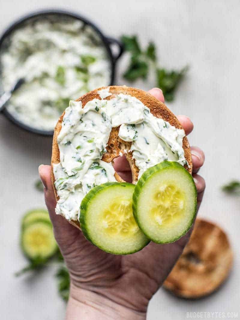 A hand holding a toasted bagel smeared with Scallion Herb Cream Cheese Spread and a couple of cucumber slices