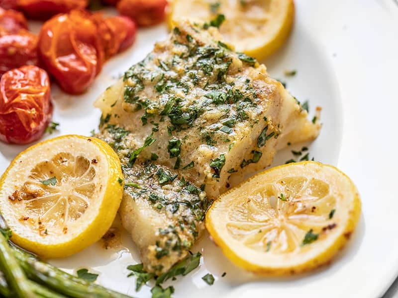 Close up front view of a piece of Garlic Butter Baked Cod on a plate with tomatoes and asparagus.