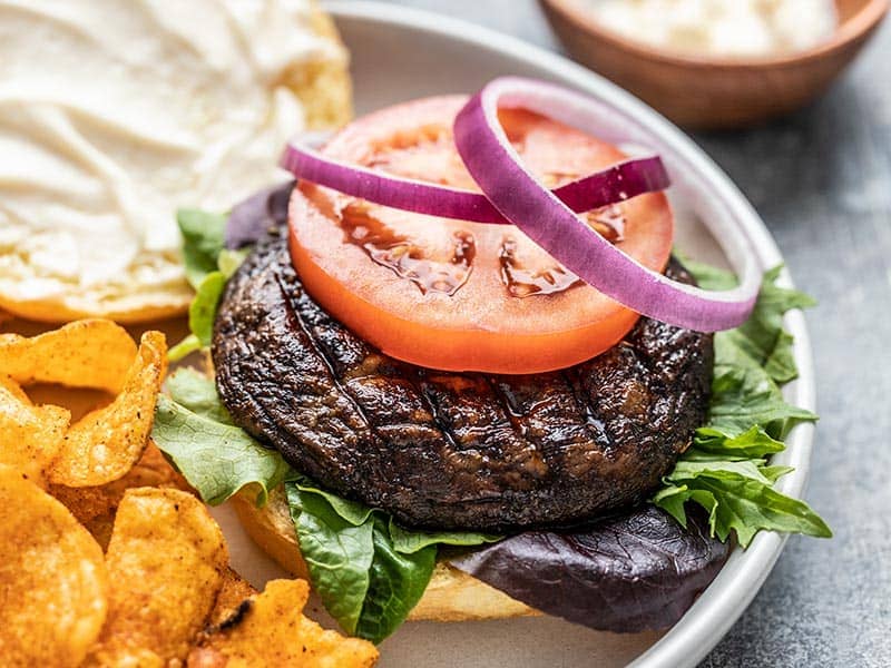 Close up side view of a marinated portobello mushroom burger topped with a slice of tomato and two red onion rings.