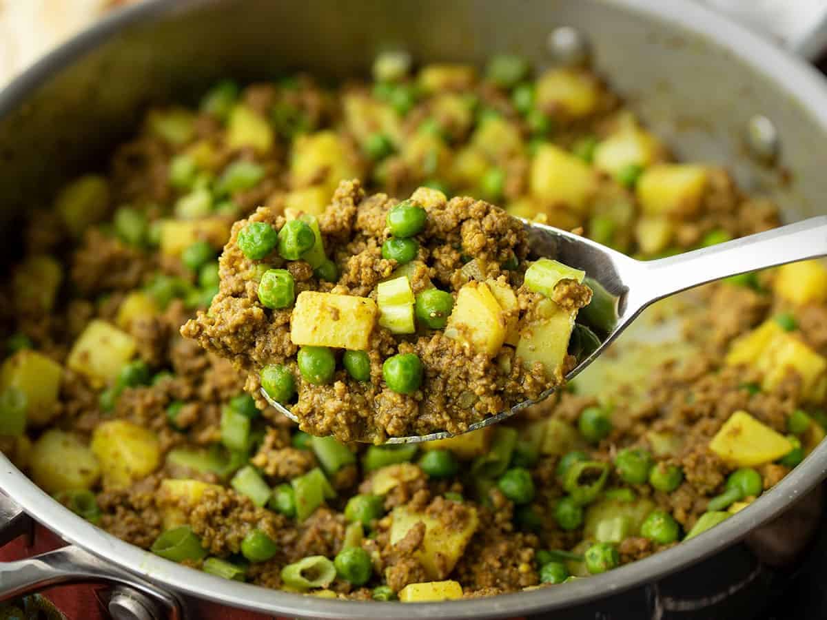 close up of a scoop of curried ground beef with peas and potatoes in front of the skillet