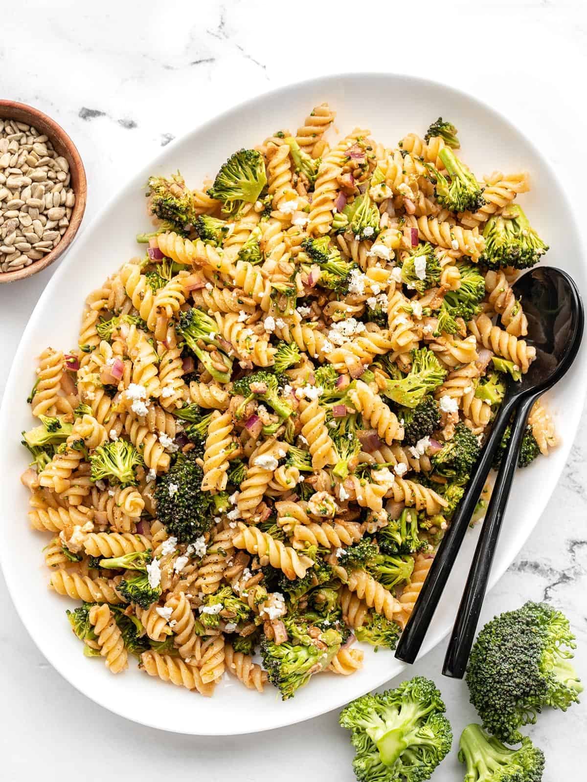 Overhead of an oval serving tray full of broccoli pasta salad with black utensils in the side