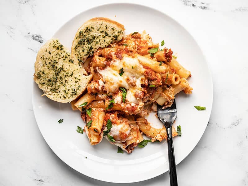A white plate with baked ziti, garlic bread, and a black fork.