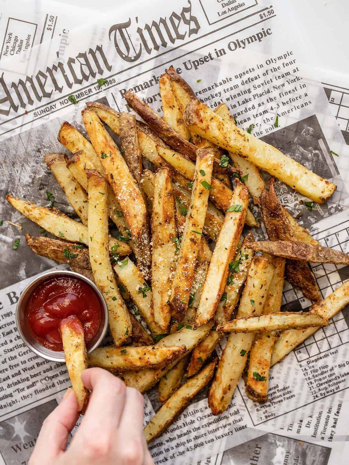 a hand dipping one garlic parmesan fry into ketchup next to a pile of fries