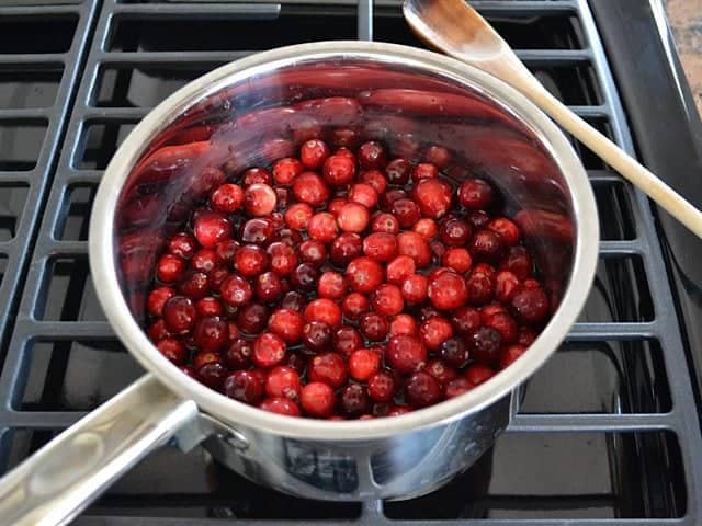 Cranberries added to boiling sugar water in pot on stove top, wooden spoon on the side 
