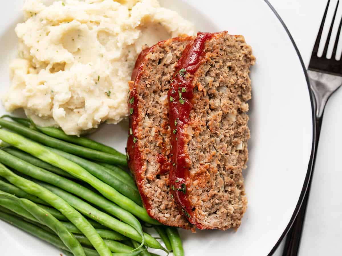 Two slices of meatloaf on a plate with mashed potatoes and green beans