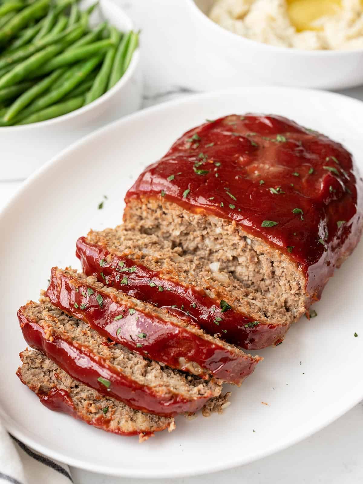 Side view of sliced meatloaf on a platter, side dishes in the background