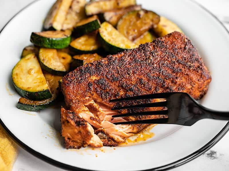 A fork flaking a piece of blackened salmon on the plate with zucchini
