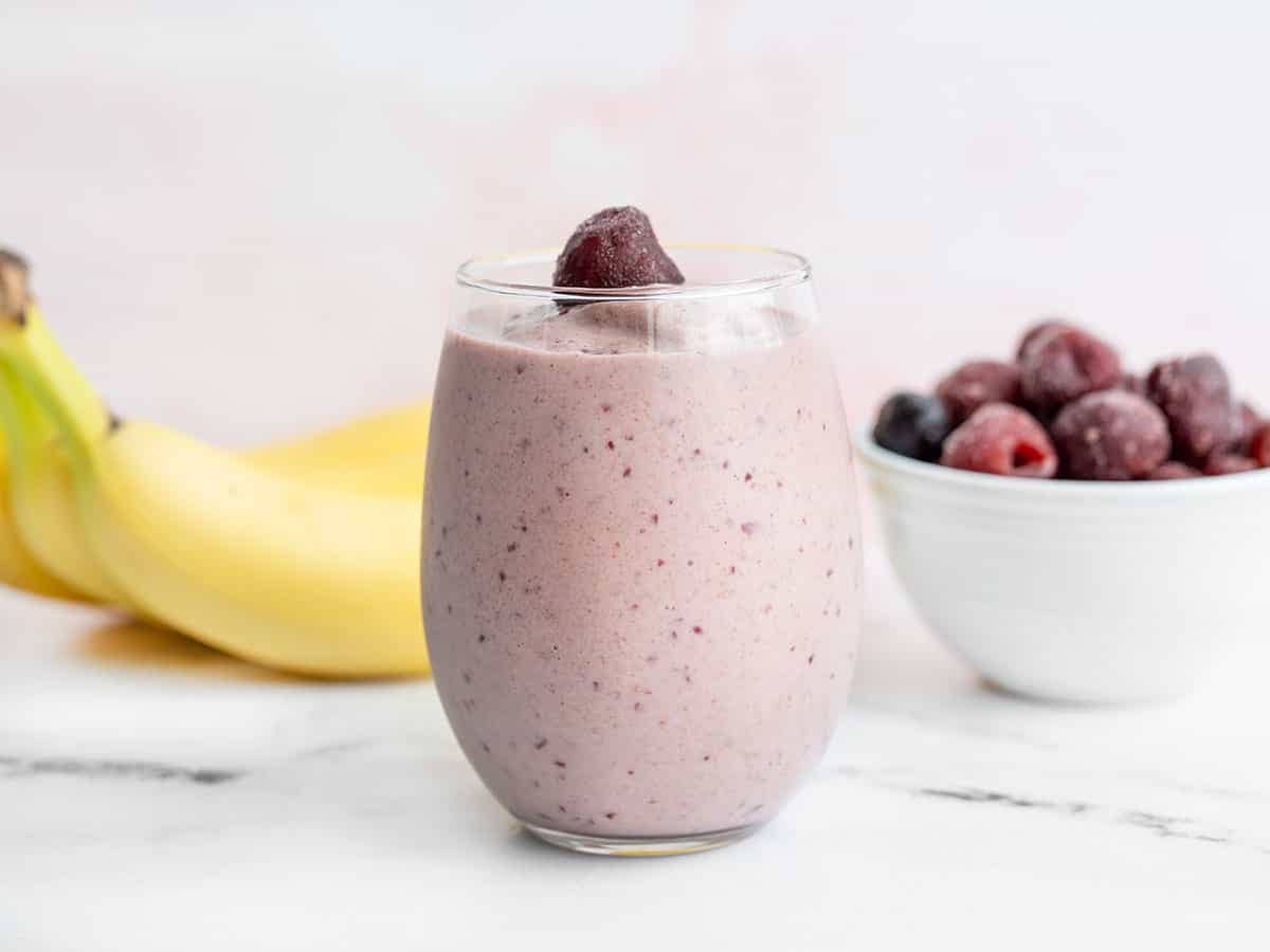 A glass filled with a cherry almond smoothie with bananas and a bowl of frozen cherries in the back