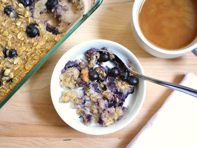 Top view of a bowl of Blueberry Banana Baked Oatmeal with whole dish of oatmeal and a cup of coffee in the background 