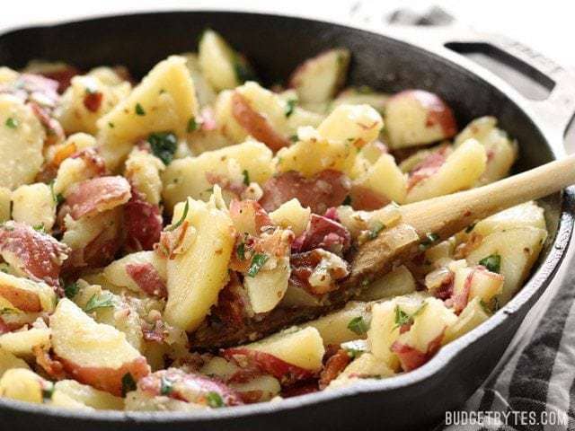 Side view of German Potato Salad in a cast iron skillet with a wooden spoon lifting a scoopful. 