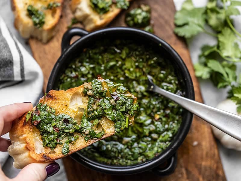 A toasty piece of French Bread being dipped into a bowl of chimichurri sauce