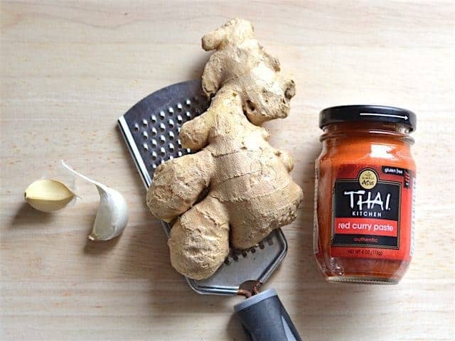 Garlic, Ginger sitting on a small-holed cheese grater, and a jar of Thai Curry Paste on a wooden cutting board