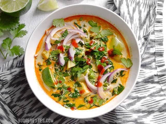 Thai Curry Vegetable Soup fully garnished with lime wedges, thinly sliced red onion, cilantro, and sriracha. 