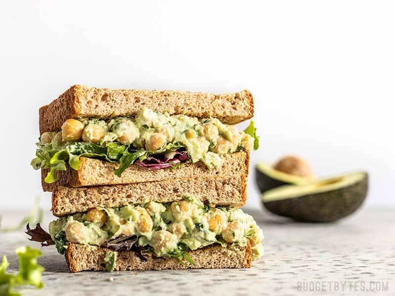 Two halves of a Scallion Herb Chickpea Salad sandwich on wheat bread, stacked. An avocado in the background. 