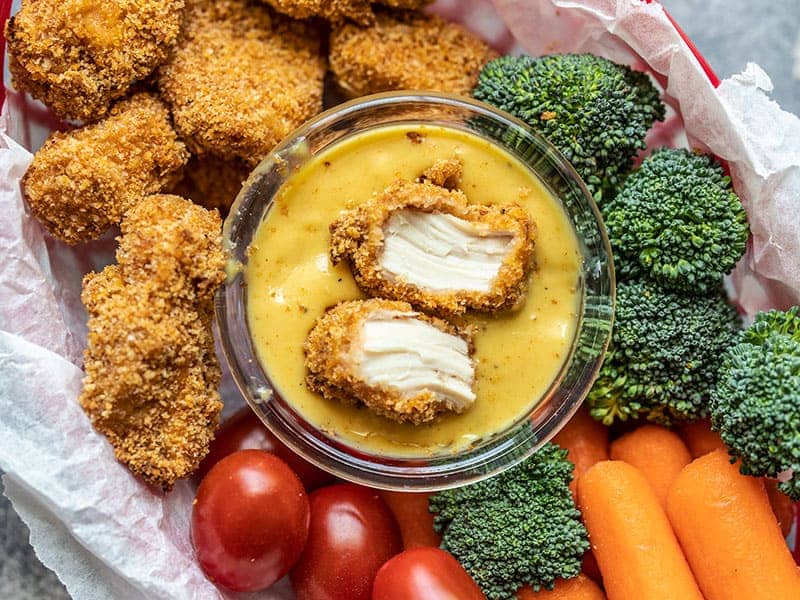 One chicken nugget ripped in half, sitting in a bowl of honey mustard, surrounded by vegetables and nuggets.
