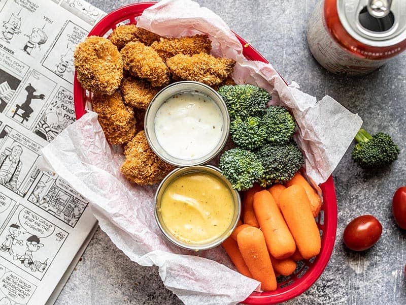 A basket full of Homemade Baked Chicken Nuggets with broccoli and carrots, and dishes of ranch and honey mustard dip.
