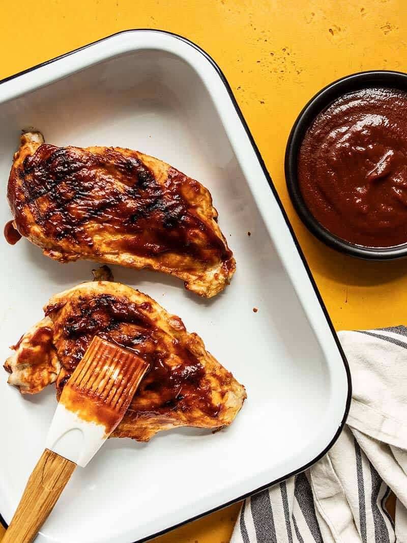 Homemade BBQ sauce being brushed onto a grilled chicken breast in a white dish with a bowl of sauce on the side.