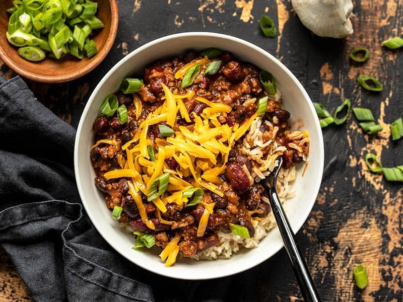 Overhead view of a bowl full of BBQ Beef and Beans, served with rice and topped with cheddar and green onion. A bowl of green onion on the side.