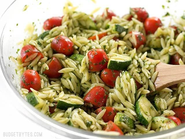 Close up side view of the bowl full of Zucchini and Orzo Salad with Chimichurri, with a wooden spoon stuck in the side