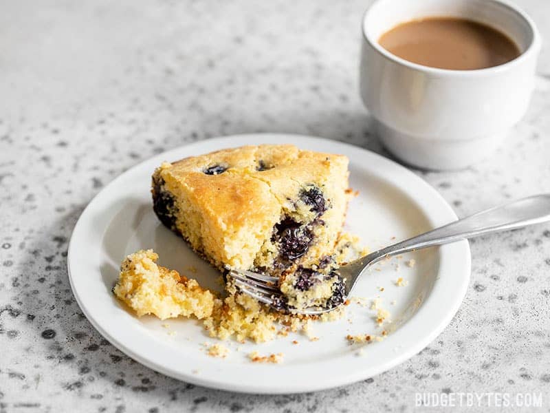 A slice of Lemon Blueberry Cornbread on a plate with a fork and cup of coffee in the background.