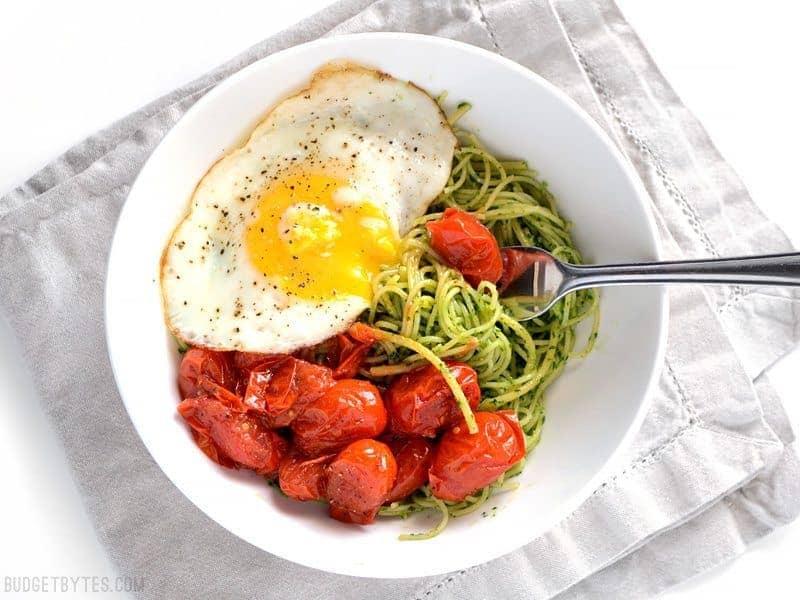 Overhead view of a bowl of parsley pesto pasta with blistered tomatoes and fried egg, yolk broken and dripping